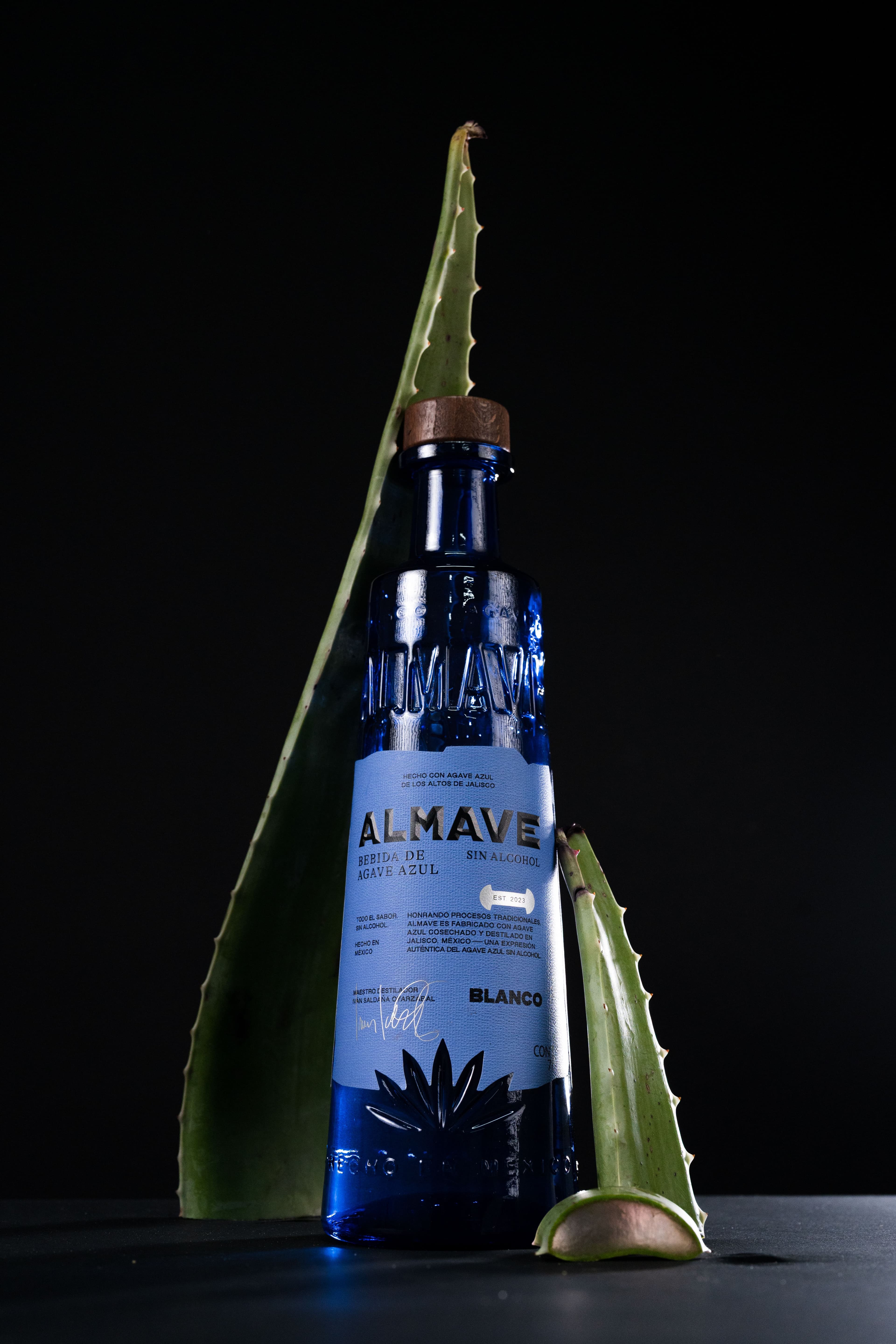 Studio shot of Almave’s premium blue bottle, crafted from blue agave, elegantly staged with agave leaves against a dark background to highlight its authentic and sophisticated design.