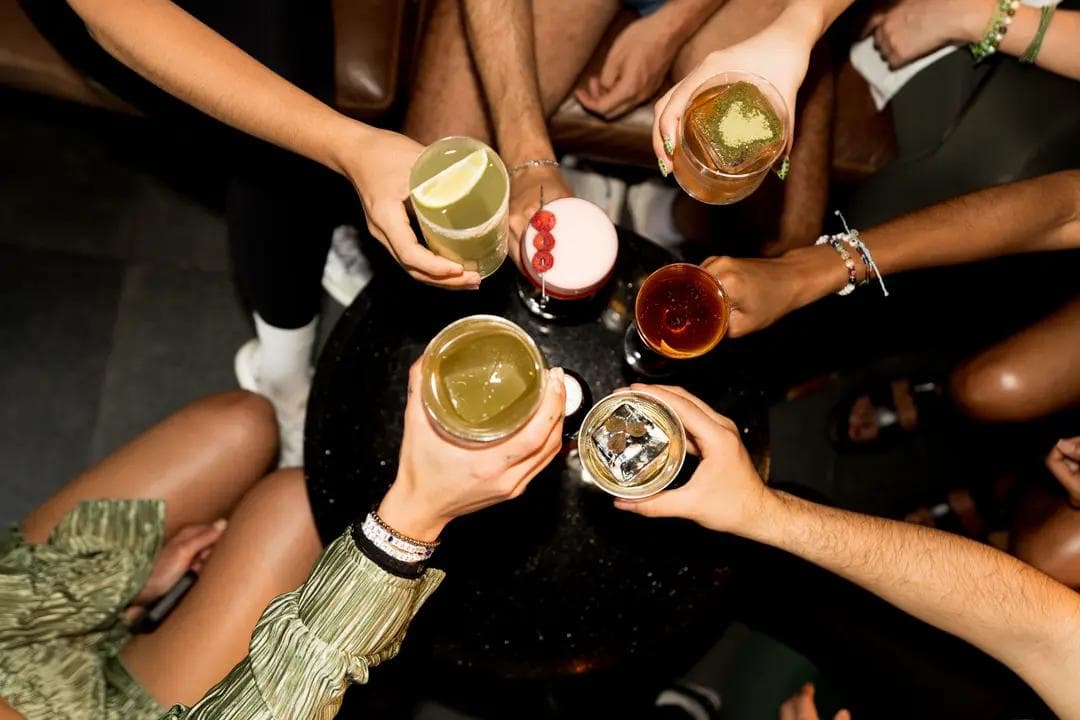 Top view of friends raising an assortment of colorful cocktails around a dark table, capturing a moment of celebration and connection with a premium, modern vibe.