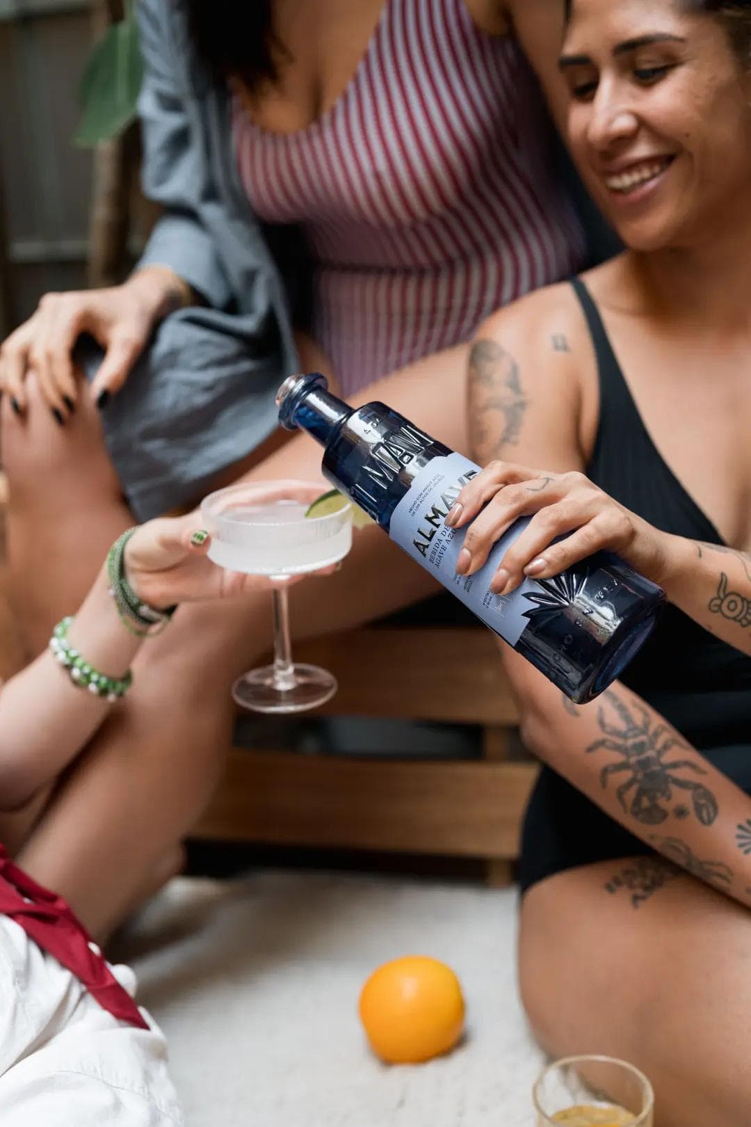 Friends enjoying drinks together; one person pours clear liquor from a blue bottle labeled 'Almana' into a cocktail glass with a lime slice, while others sit nearby in swimwear, smiling and relaxing.